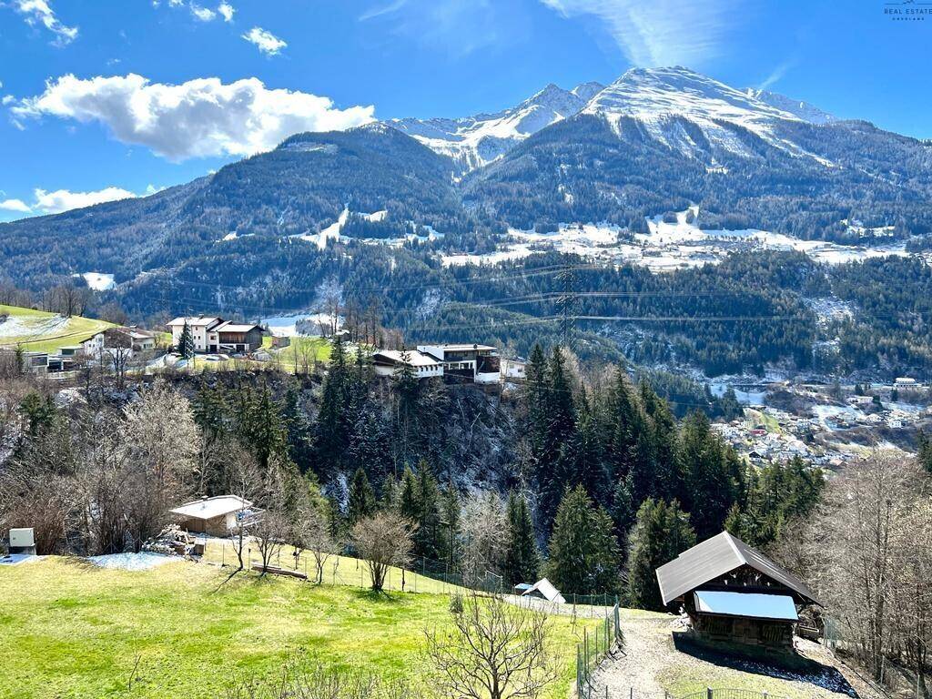 Weitläufige Berglandschaft mit grünen Wiesen, vereinzelten Häusern und schneebedeckten Gipfeln.