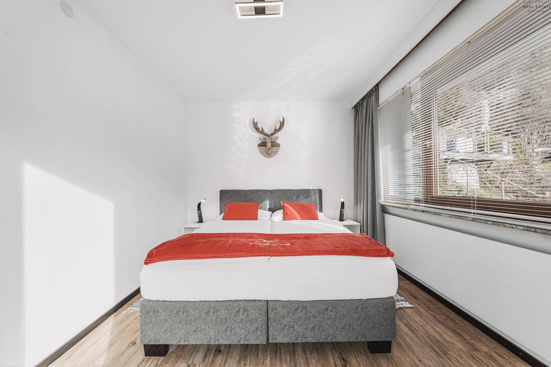 Helles Schlafzimmer mit Doppelbett, Holzboden und Fenster mit Jalousien, dekoriert mit Hirschgeweih.