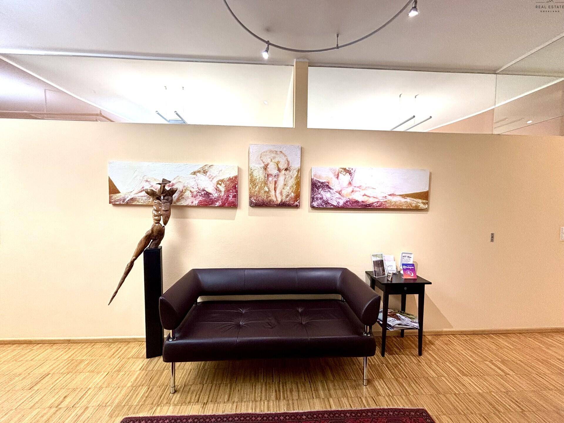Wartebereich mit dunklem Ledersofa, Beistelltisch und Kunstwerken an einer beigen Wand.