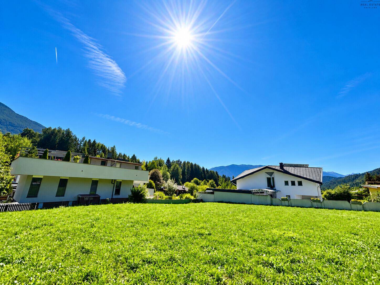 Weite Grünfläche mit strahlendem Sonnenschein, Bergen und Häusern im Hintergrund.