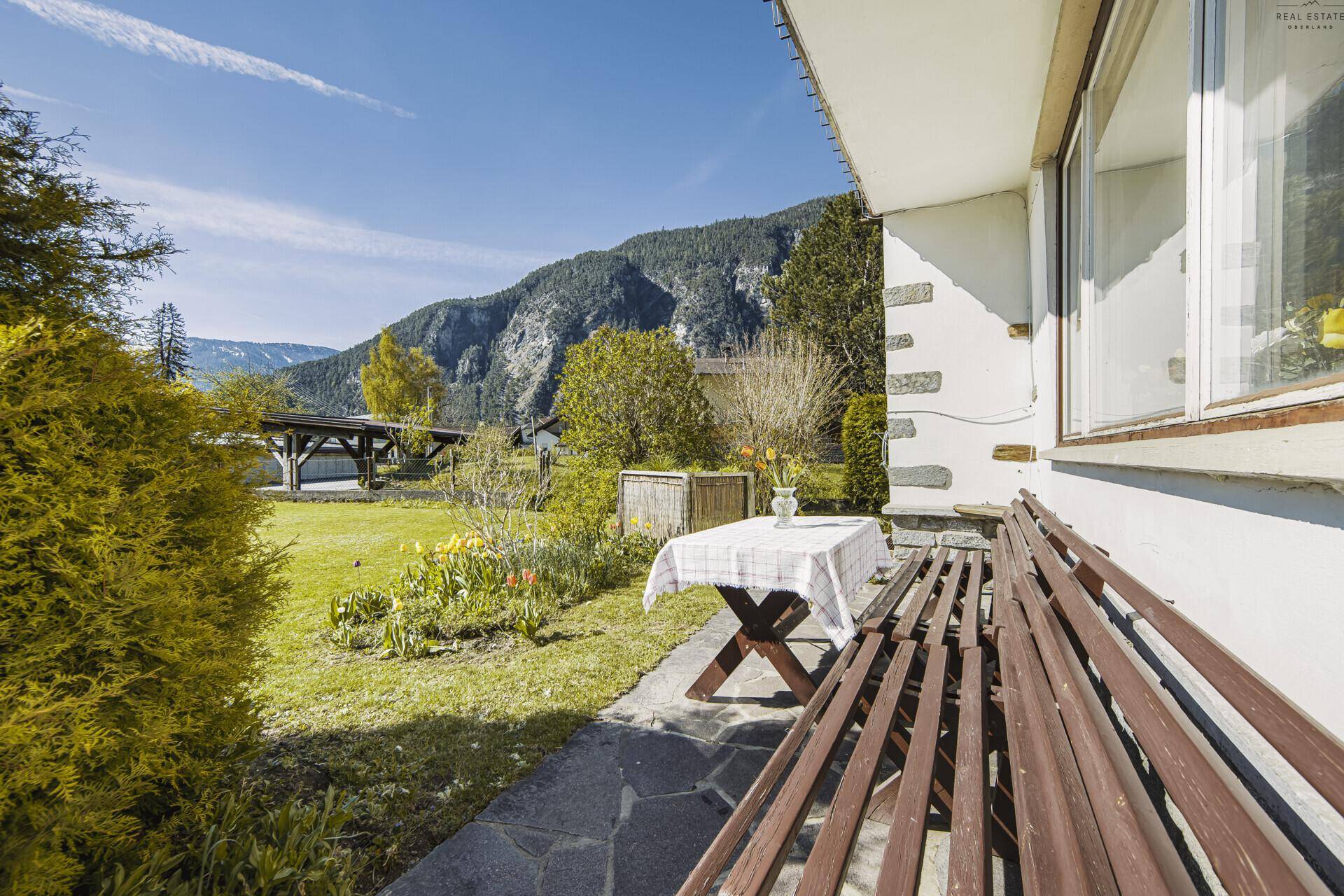 Gemütlicher Sitzbereich im Freien mit Holztisch und Bank, umgeben von Grün und Bergblick.