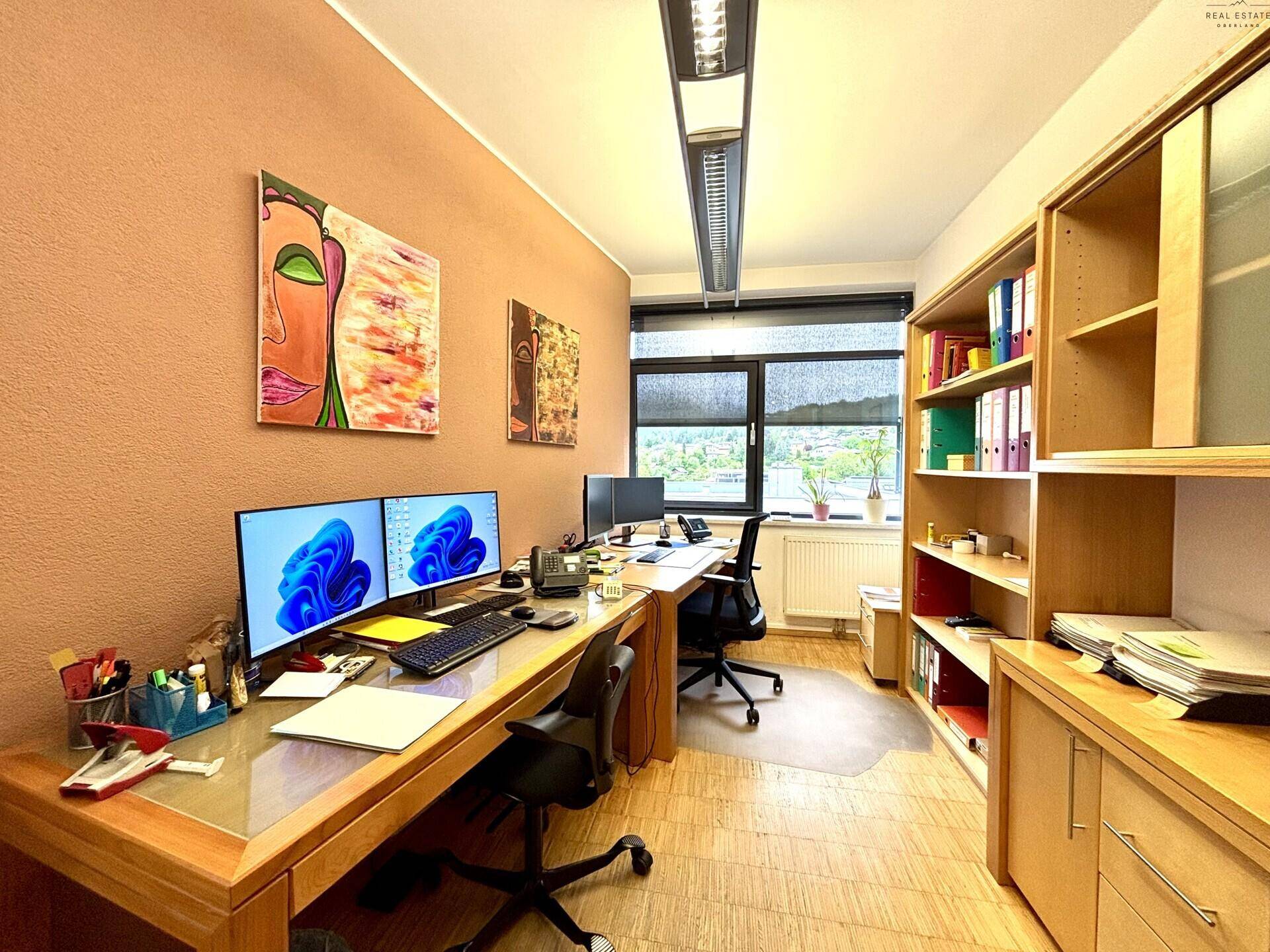 Bürozimmer mit zwei Arbeitsplätzen, Computern, Holzregalen und Fenstern mit Ausblick.