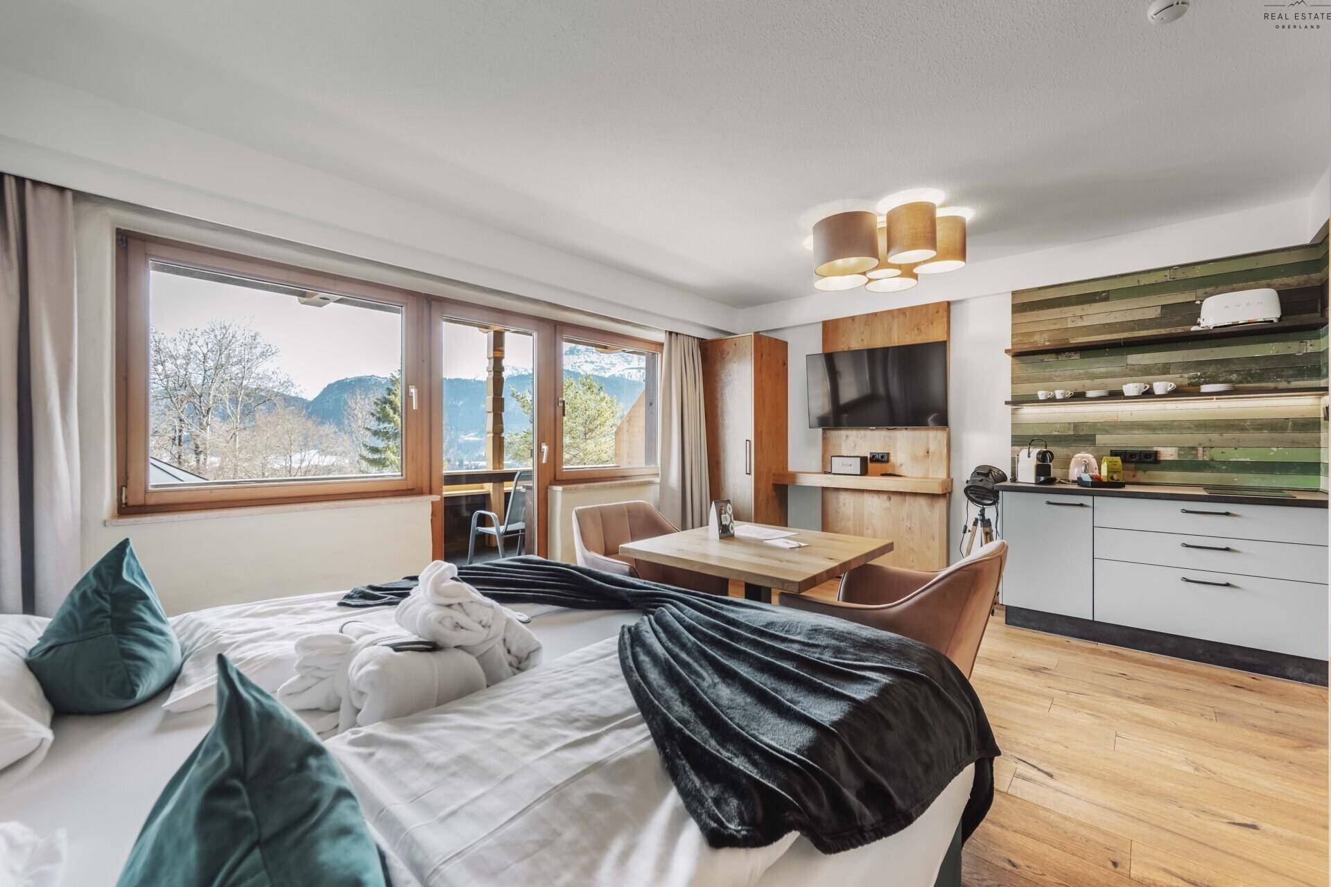 Helles Schlafzimmer mit Doppelbett, kleiner Küchenzeile und großzügigen Fenstern mit Bergblick.