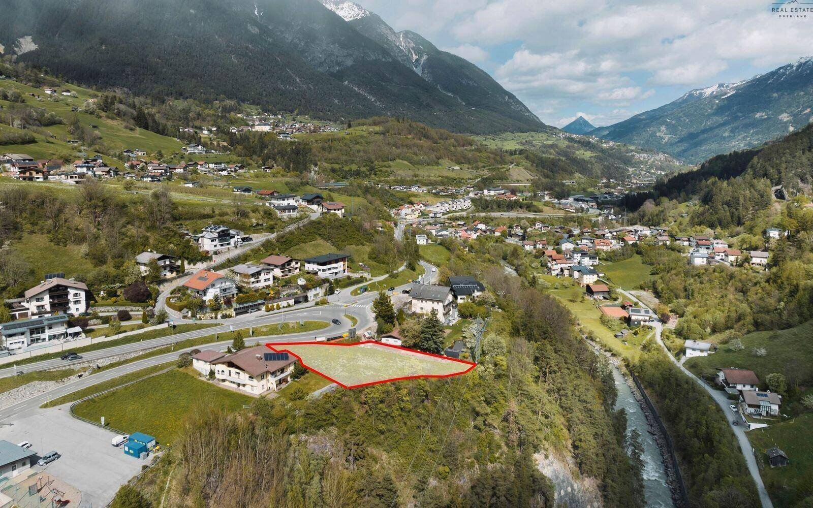 Panoramische Luftaufnahme eines Baugrundstücks in einem alpinen Tal mit umliegenden Häusern und Bergen.
