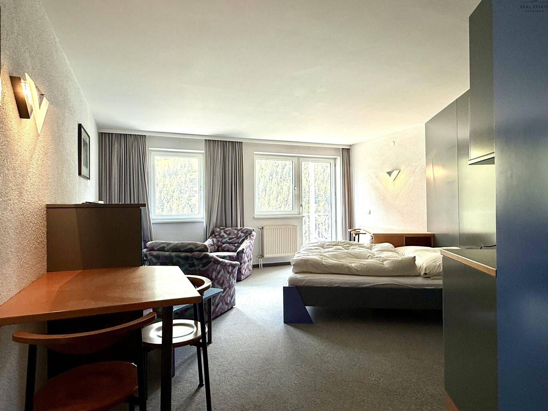 Schlafzimmer mit Bett, kleinem Esstisch und Blick auf die Umgebung durch große Fenster.