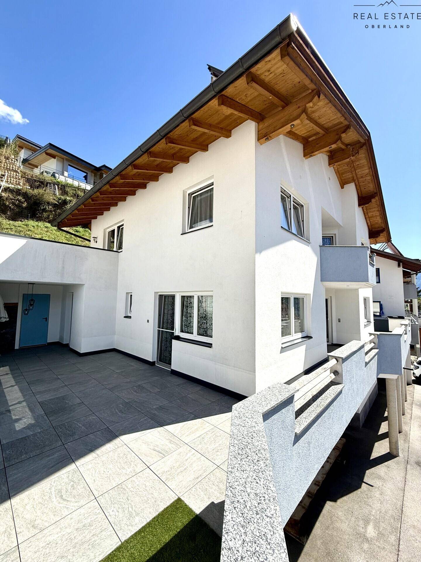 Außenansicht des Hauses mit weißer Fassade, Holzdachüberstand und großer gefliester Terrasse.
