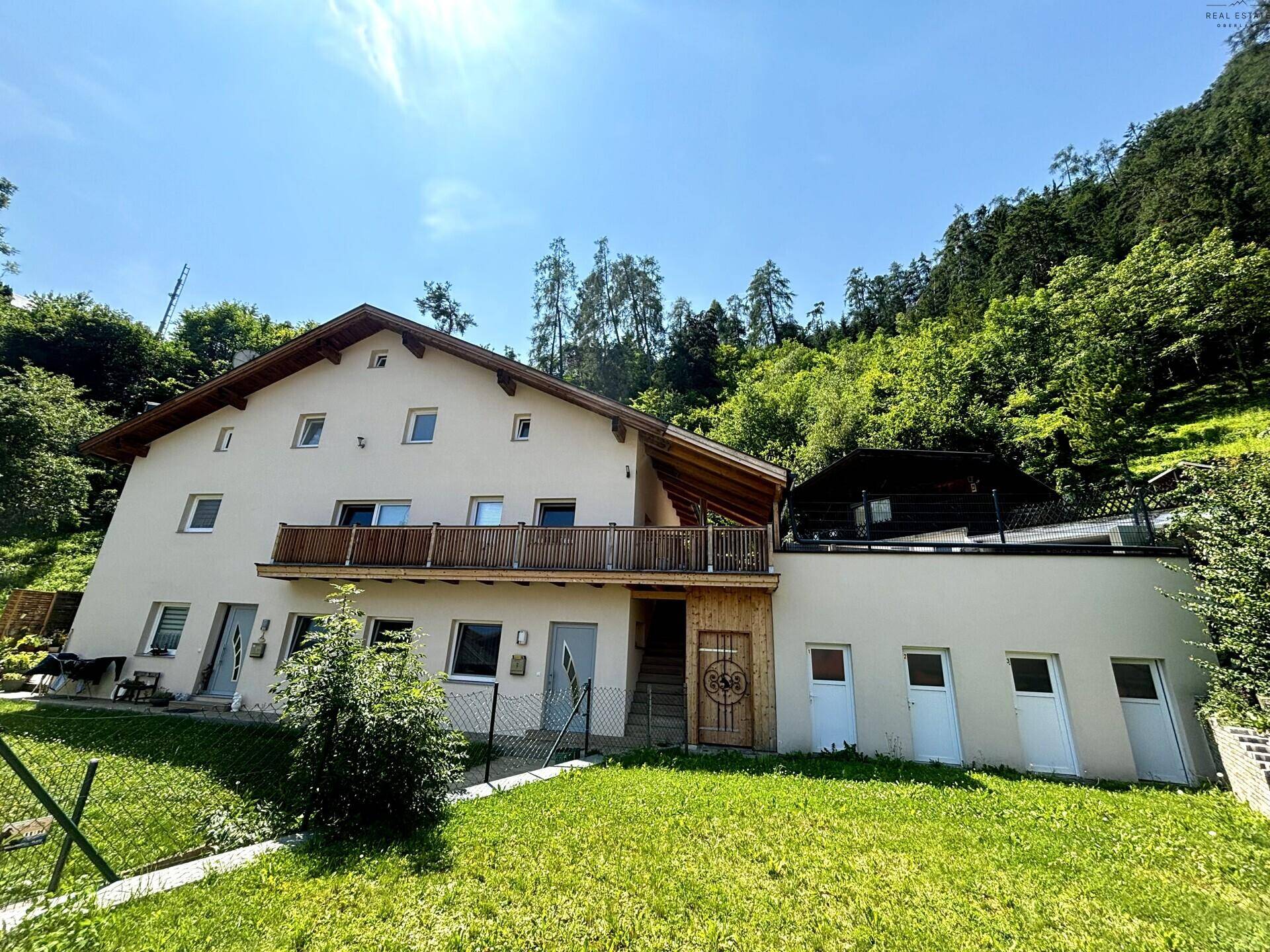 Ansicht des Hauses mit Garten und angrenzendem Hang in ländlicher Umgebung unter blauem Himmel.