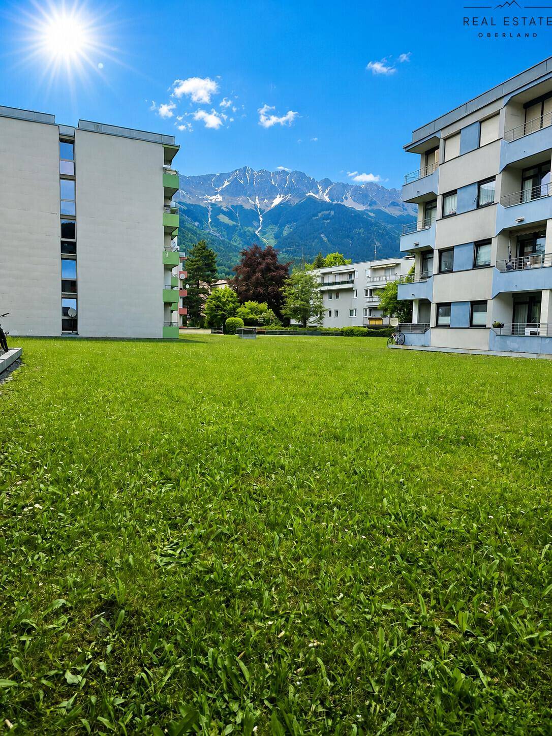 Grüne Rasenfläche vor Wohngebäuden mit beeindruckendem Bergblick unter blauem Himmel.