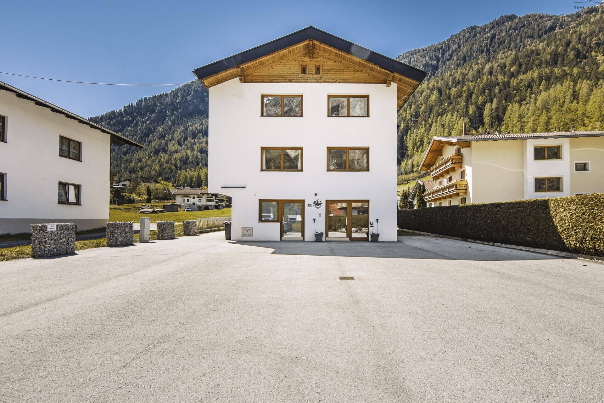 Weißes Wohnhaus mit Holzelementen und großem Vorplatz, umgeben von alpiner Landschaft.