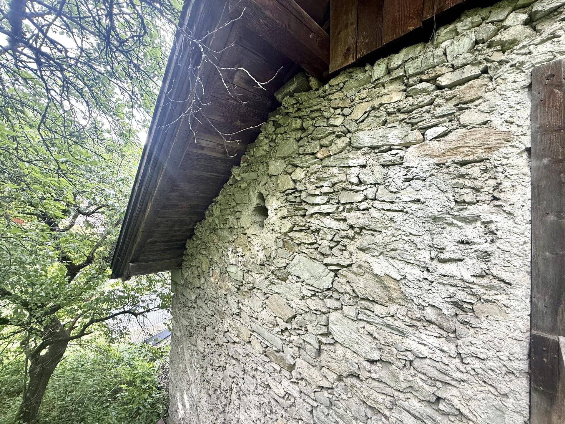Rustikale Steinmauer eines alten Gebäudes, umgeben von üppigem Grün und Bäumen.