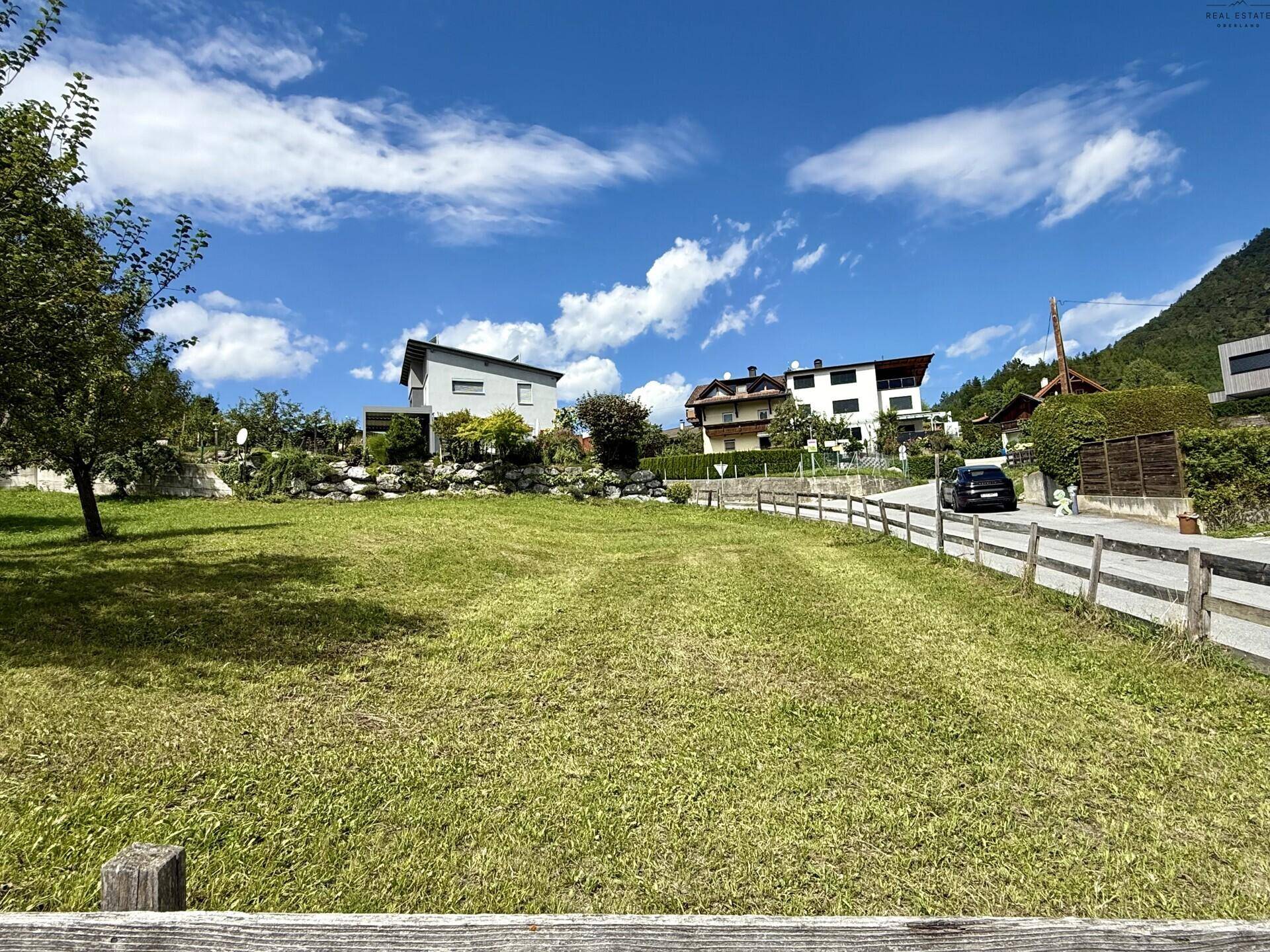 Weitläufige grüne Fläche mit Häusern und einer Bergkulisse unter einem klaren blauen Himmel.