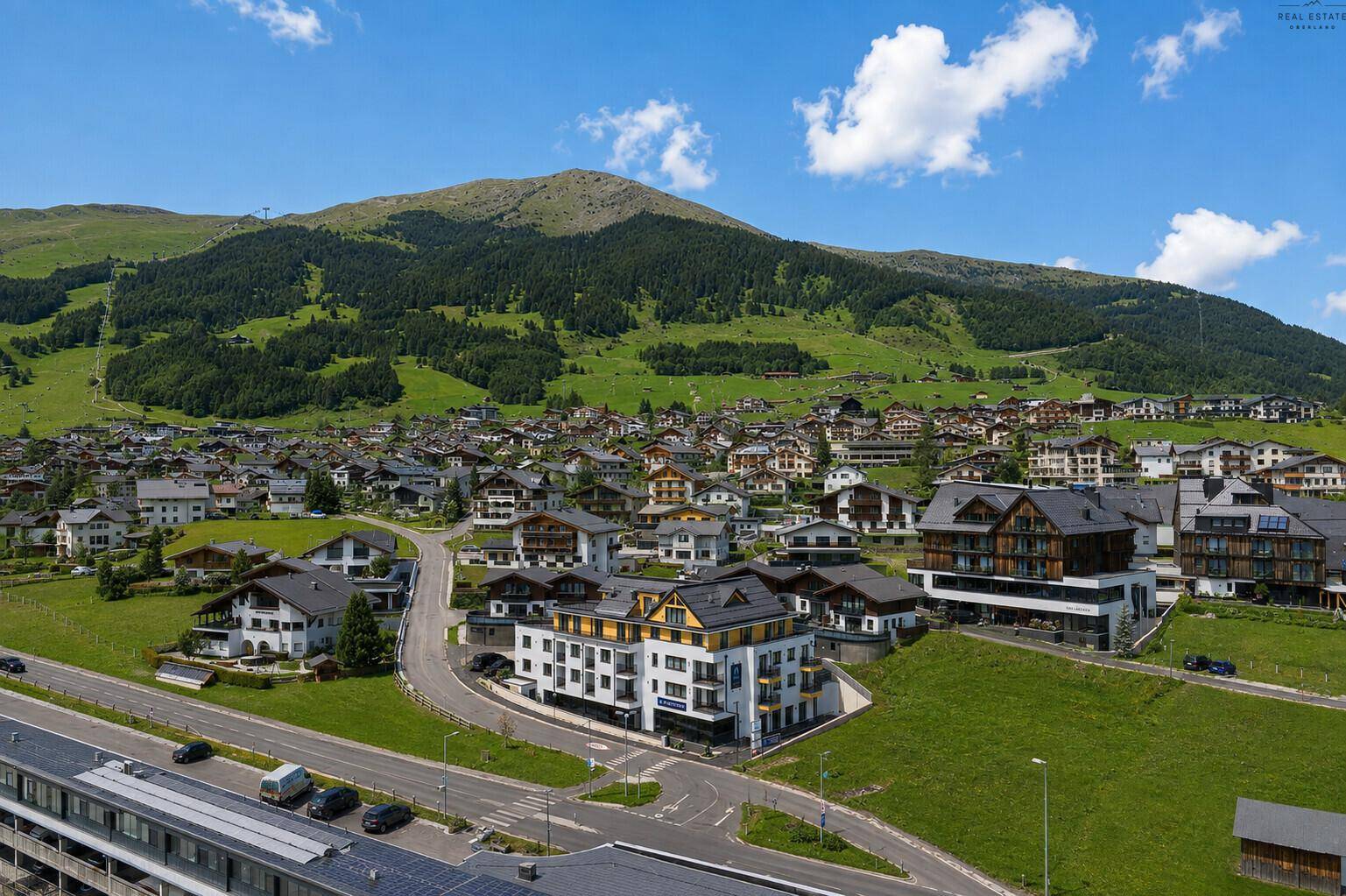 Weitläufiger Blick auf eine alpine Siedlung mit Bergen und grünen Hängen unter blauem Himmel.
