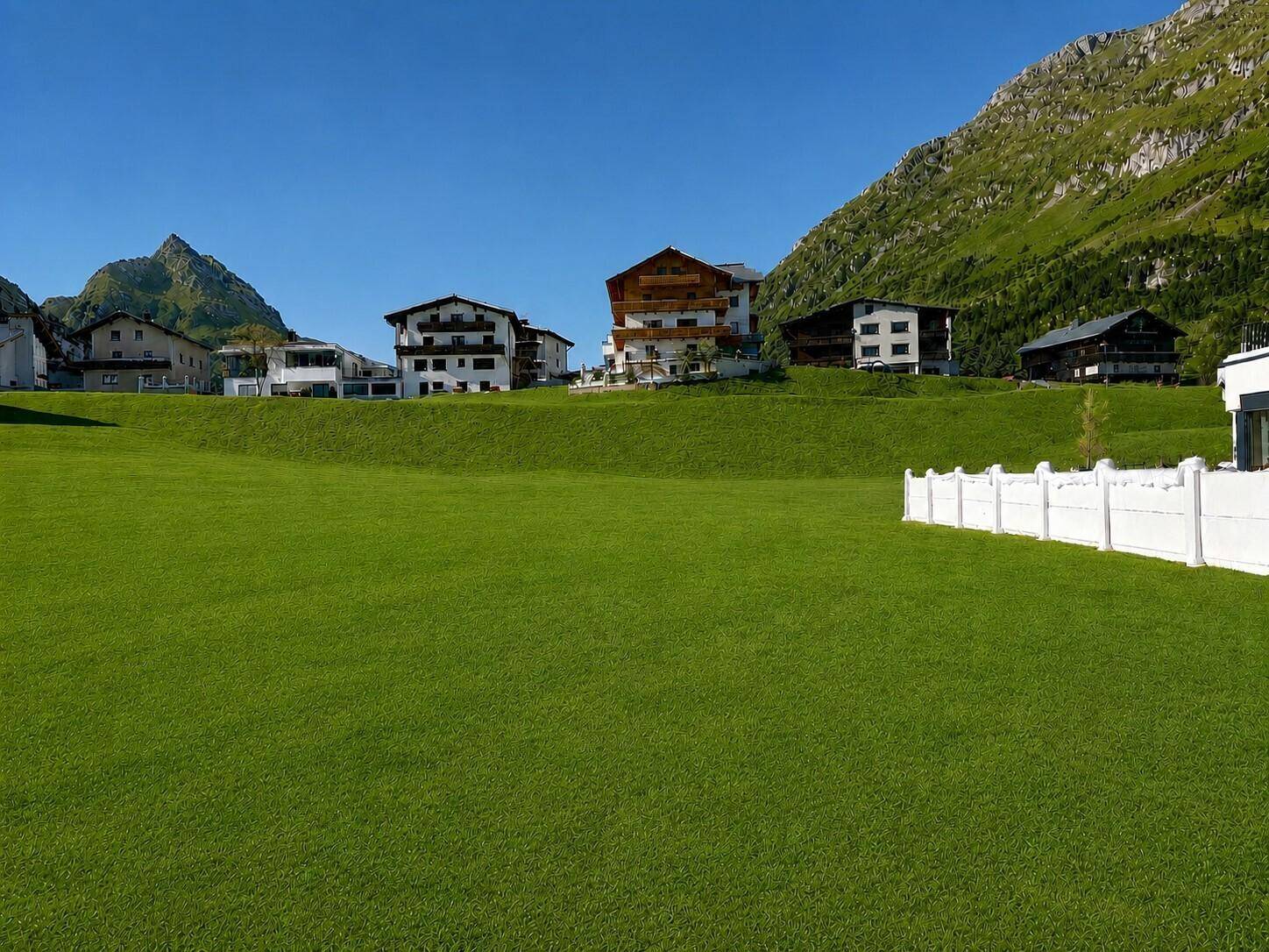 Weitläufige grüne Wiese mit weißem Zaun, verschiedenen Gebäuden und imposantem Bergpanorama unter blauem Himmel.