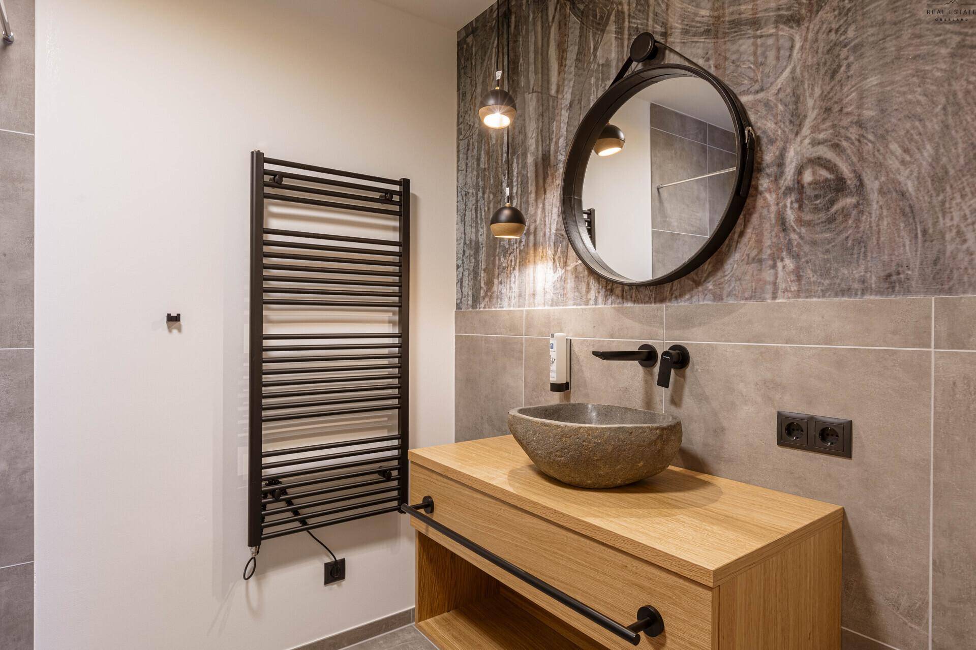 Modernes Badezimmer mit Steinswaschbecken, rundem Spiegel und beheiztem Handtuchhalter.
