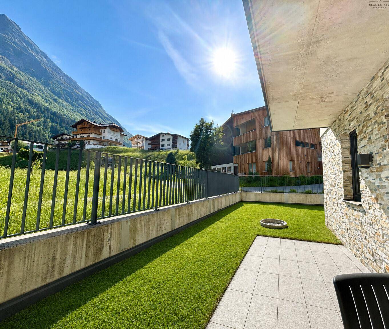 Sonnige Terrasse mit Rasen und Steinmauer, umgeben von grünen Wiesen und Bergen.