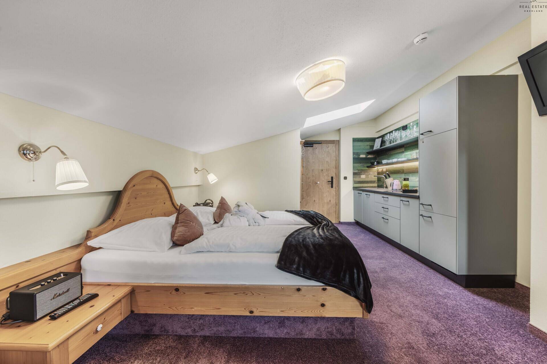 Schlafzimmer mit Holzbett, integrierter Küchenzeile und lila Teppichboden.