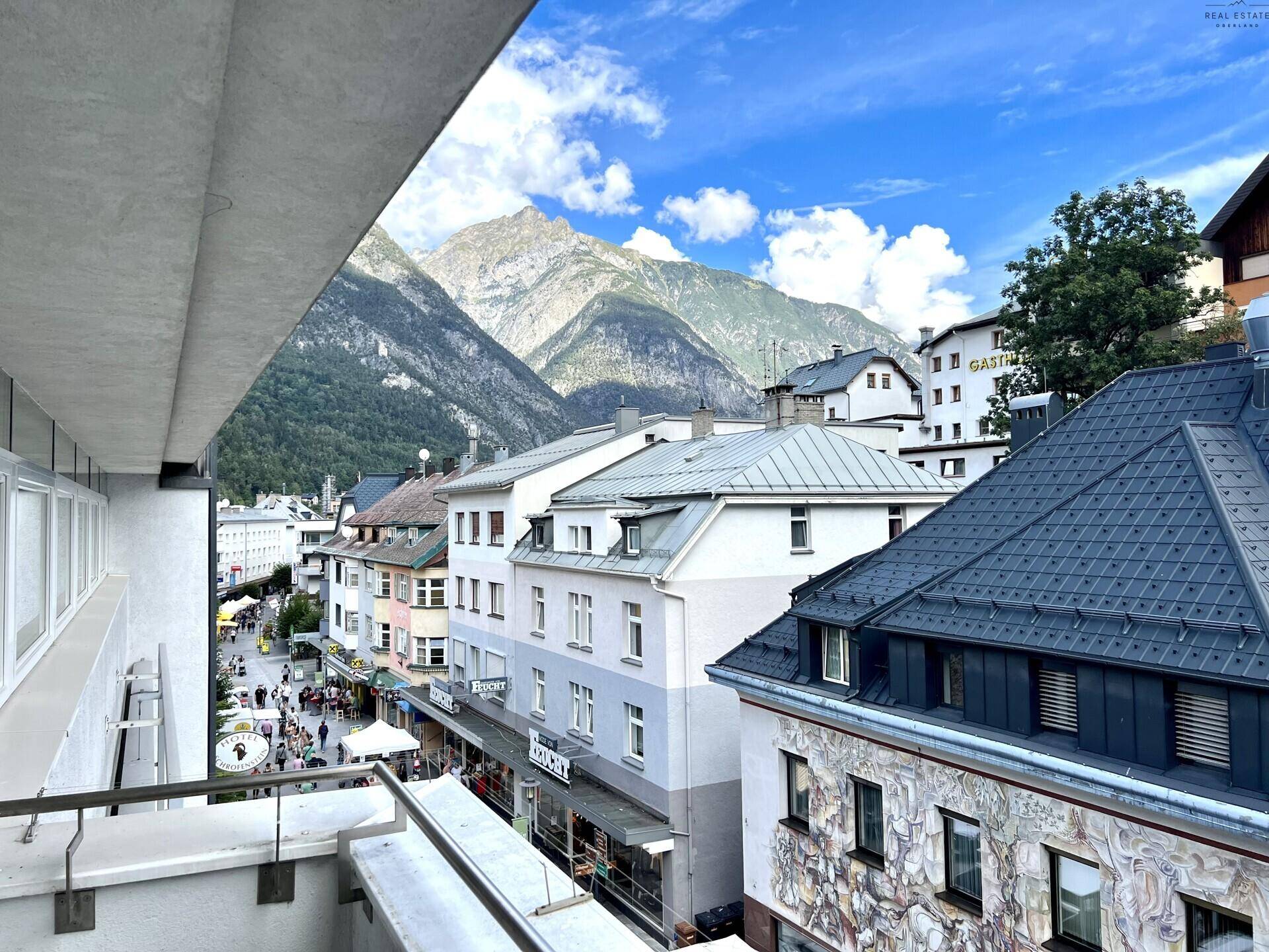 Balkon mit Blick auf eine belebte Straße, umliegende Gebäude und majestätische Berge im Hintergrund.