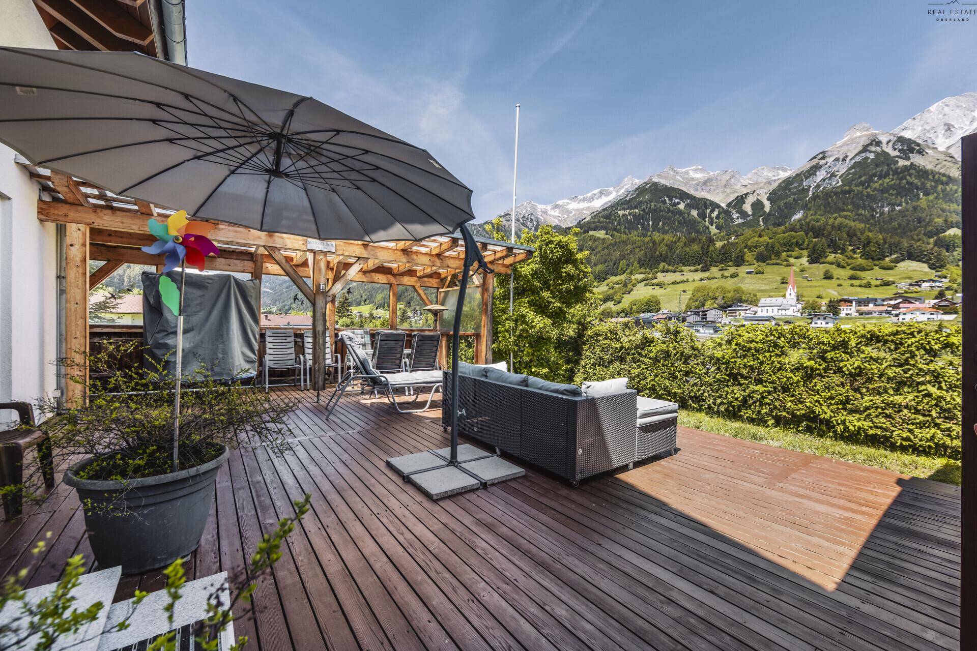 Sonnige Holzterrasse mit Lounge-Möbeln und großem Sonnenschirm, mit weitem Bergblick.