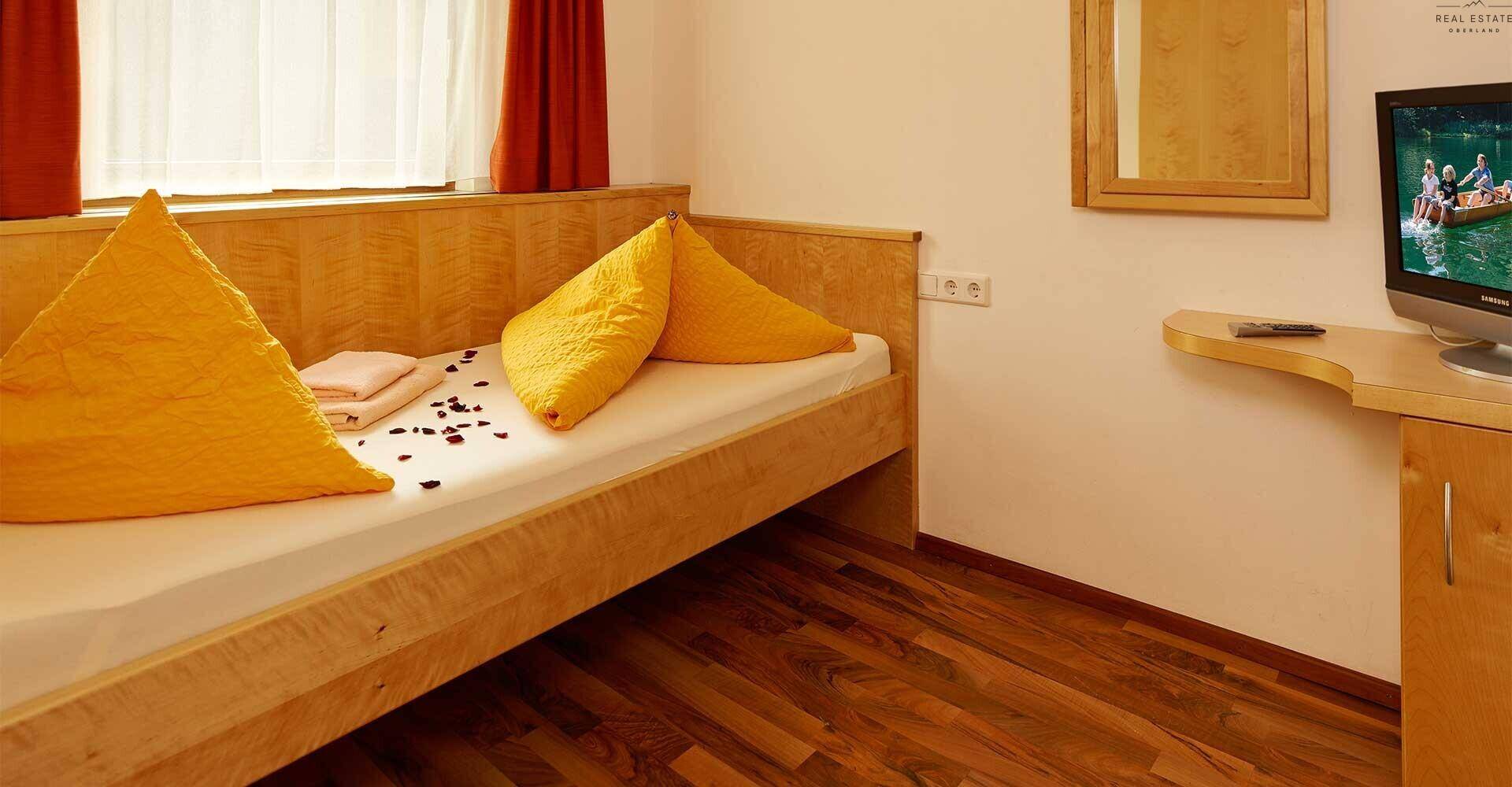 Schlafzimmer mit Einzelbett, gelben Kissen und Holzboden, ausgestattet mit einem Fernseher.