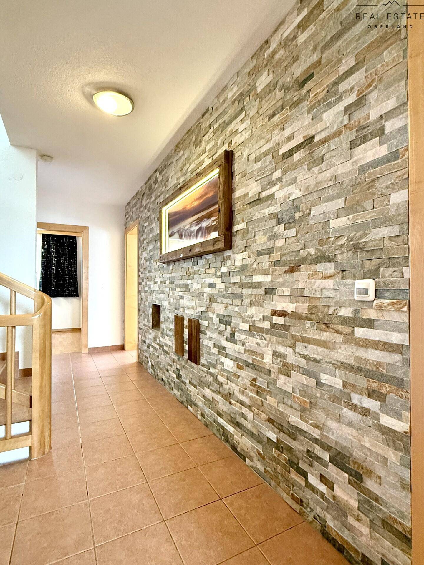 Heller Flur mit dekorativer Steinwand, Holztreppe und Terrakotta-Fliesenboden.