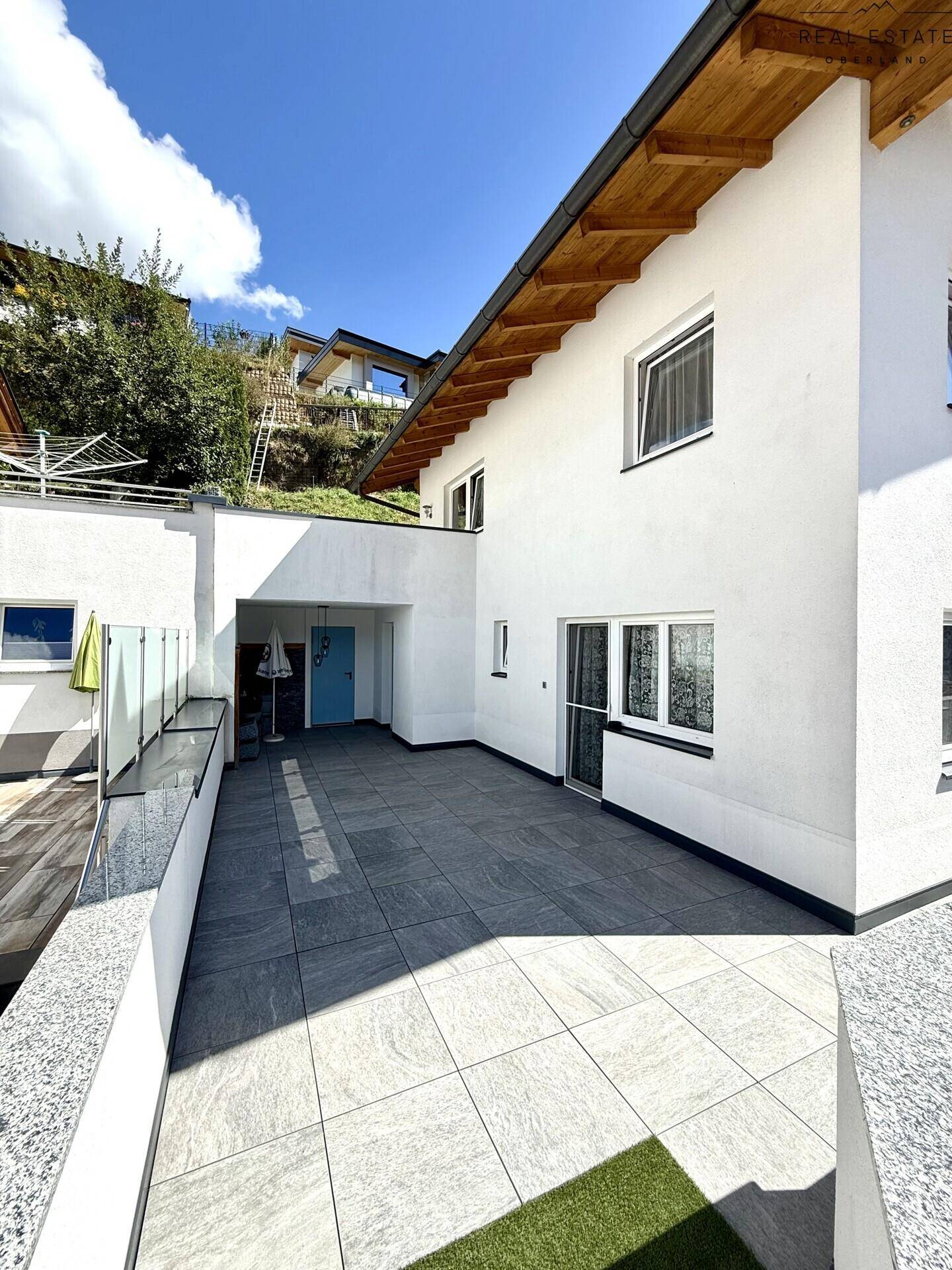 Moderne Hausfassade mit weißem Putz, Holzdach und weitläufiger, gefliester Terrasse.