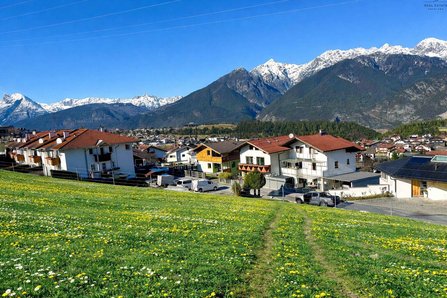 Malerische Aussicht über eine blühende Wiese auf eine Siedlung vor imposanter Bergkulisse.