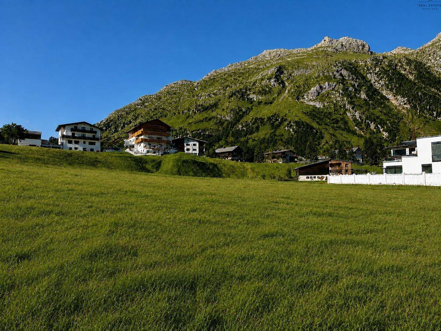 Sonnige grüne Landschaft mit verschiedenen Gebäuden und felsigen Bergen unter klarem Himmel in alpiner Region.