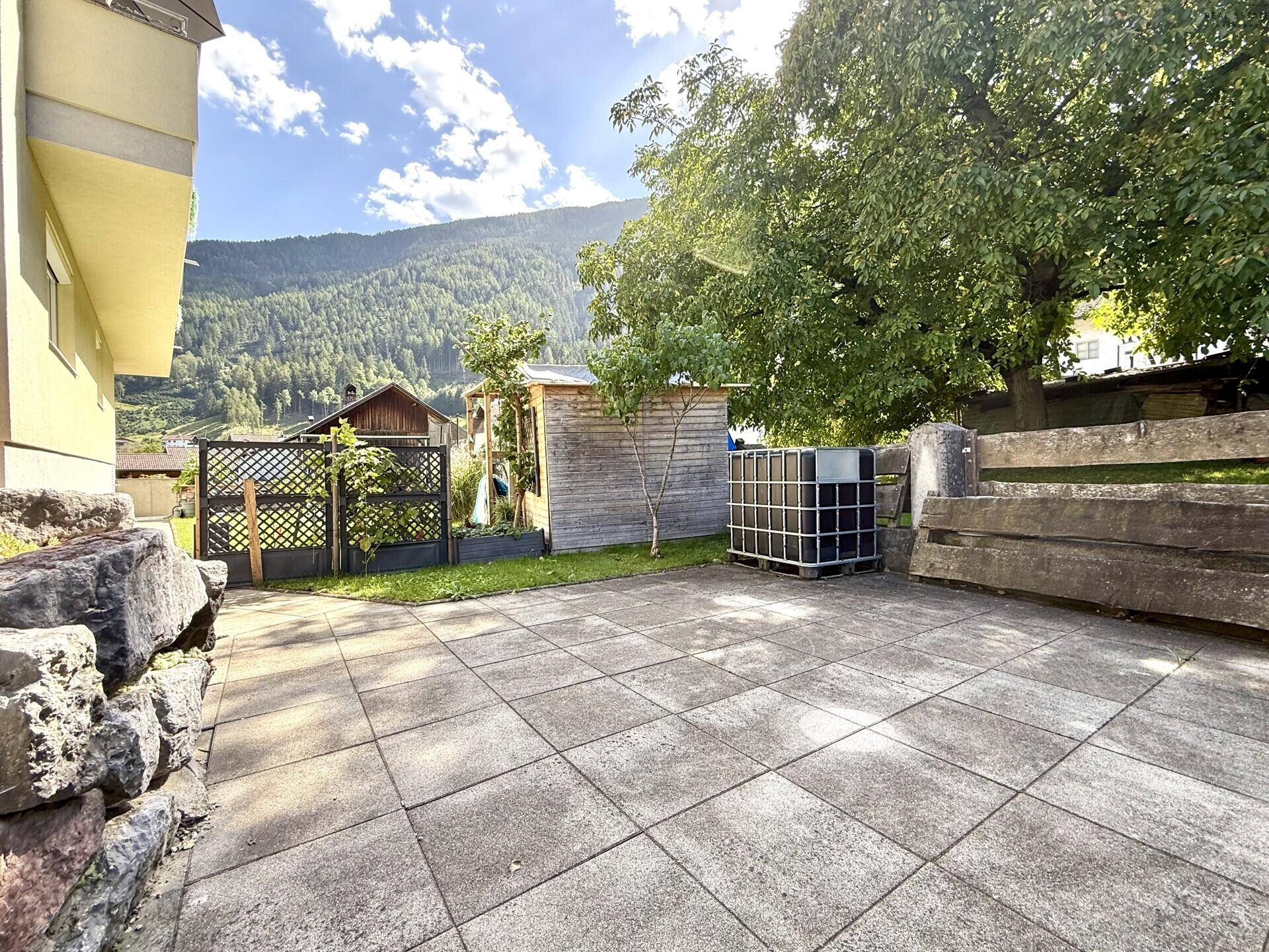 Gepflasterter Außenbereich mit Gartenhaus, Grünflächen und Blick auf die umliegende Natur.