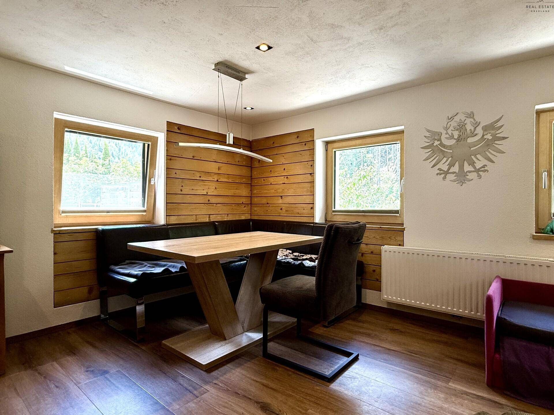 Essbereich mit Holztisch, Eckbank und Holzwandverkleidung, beleuchtet durch zwei Fenster.