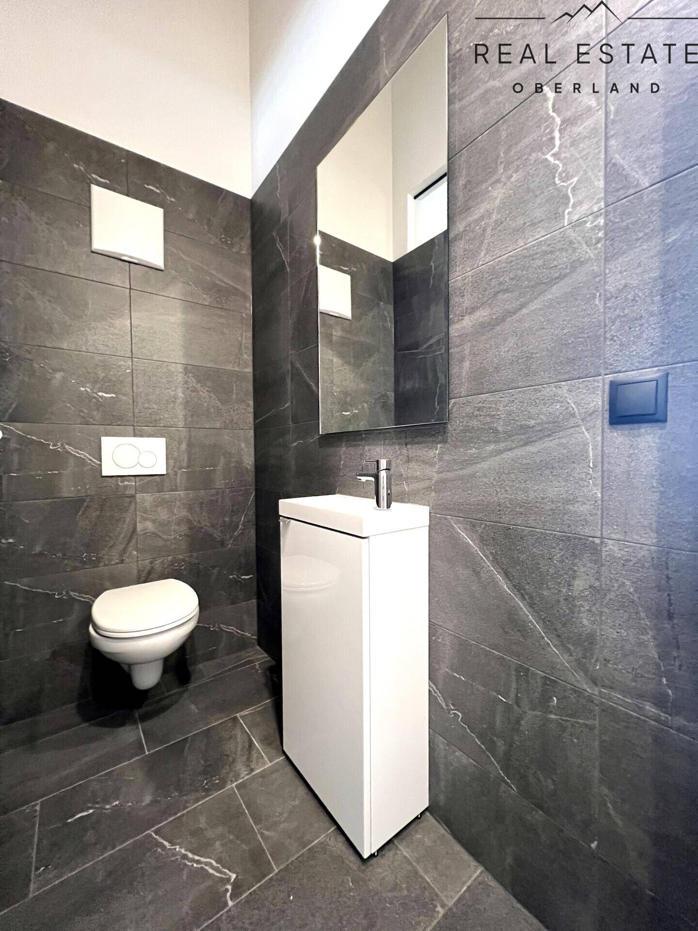 Modernes Gäste-WC mit dunklen Fliesen, wandhängendem WC und elegantem Waschtisch.