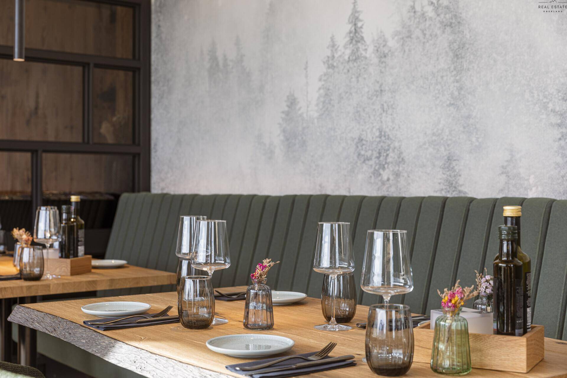 Gedeckter Holztisch mit Gläsern und Dekoration, vor einer Wand mit Waldmotiv, die eine natürliche Stimmung erzeugt.
