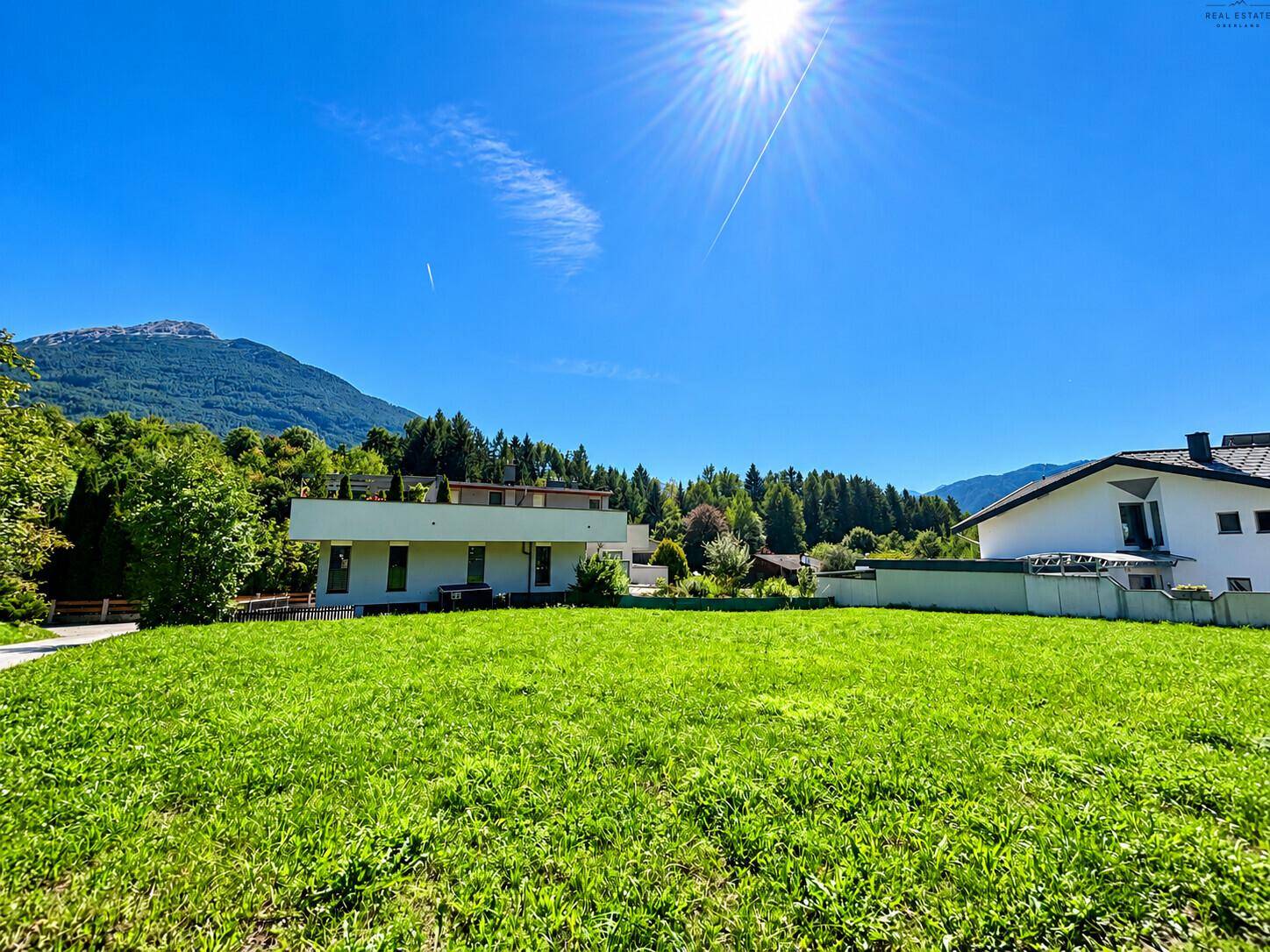 Helle Wiese mit Bergblick und Sonnenschein, umgeben von Bäumen und Häusern.
