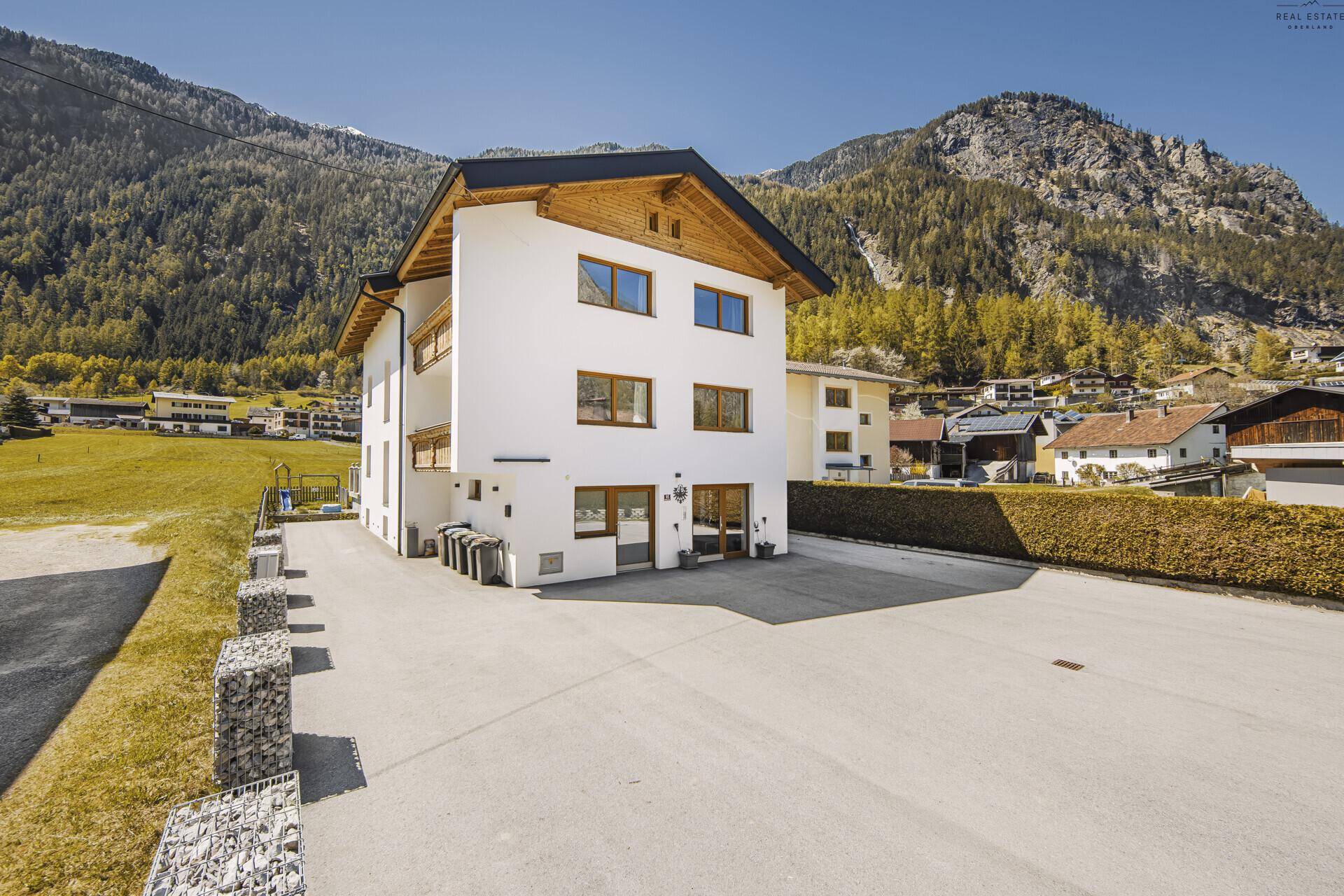 Modernes Wohnhaus mit großem Vorplatz und Hecke, umgeben von alpiner Landschaft.