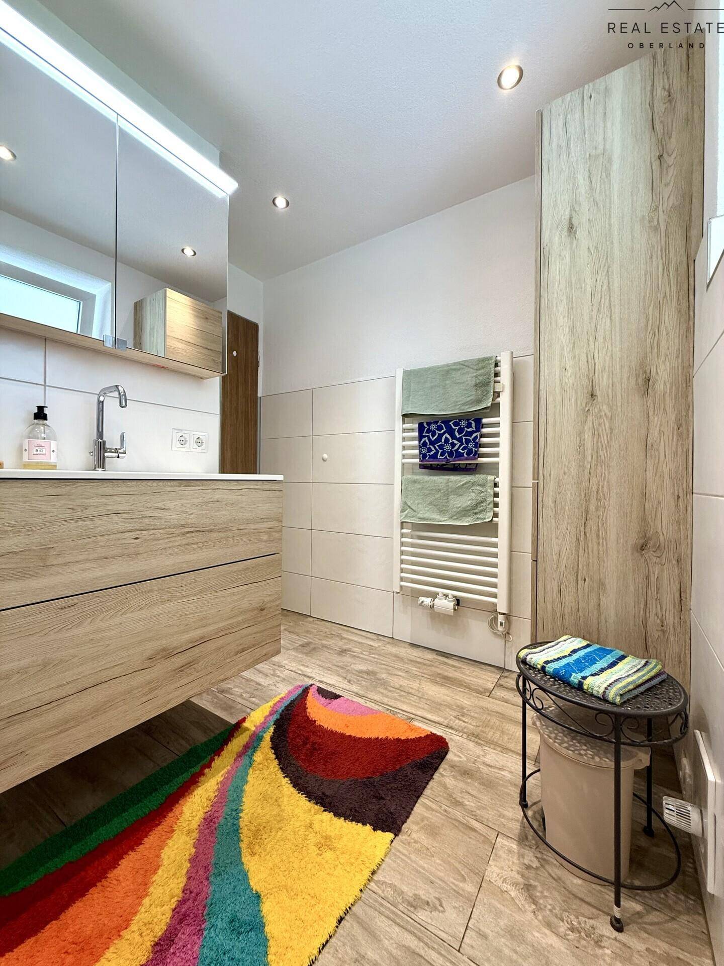 Modernes Badezimmer mit Holzwaschtisch, beheiztem Handtuchhalter und farbenfrohem Teppich.