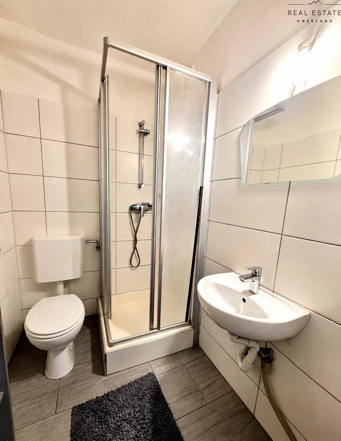 Funktionales Badezimmer mit weißen Fliesen, Duschkabine, WC und Waschbecken.