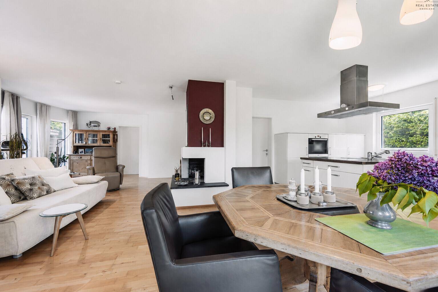 Heller offener Wohn-Essbereich mit Kamin, Holzboden und moderner Küche im Hintergrund.