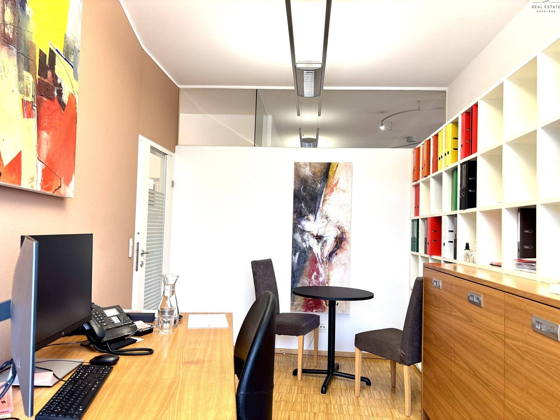 Bürobereich mit Schreibtisch, Computer, kleinem runden Tisch und weißen Regalen.