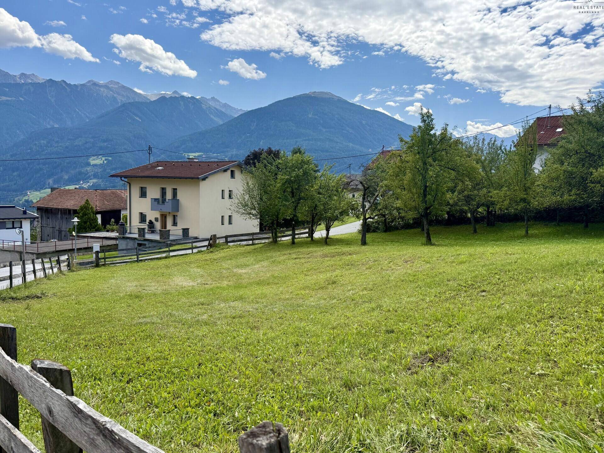 Weitläufige grüne Wiese vor einem Haus mit Bergblick unter blauem Himmel.
