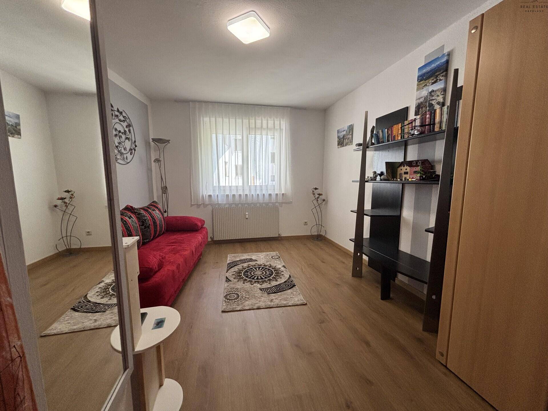 Helles Zimmer mit rotem Sofa, Bücherregal und Laminatboden, ideal als Gäste- oder Arbeitszimmer.