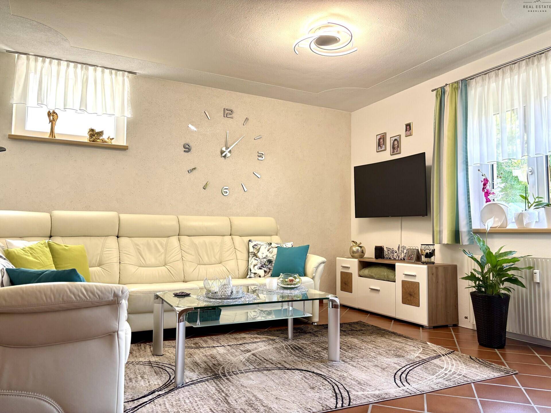 Gemütliches Wohnzimmer mit großem Sofa, Wanduhr und Blick auf den Garten durch die Fenster.