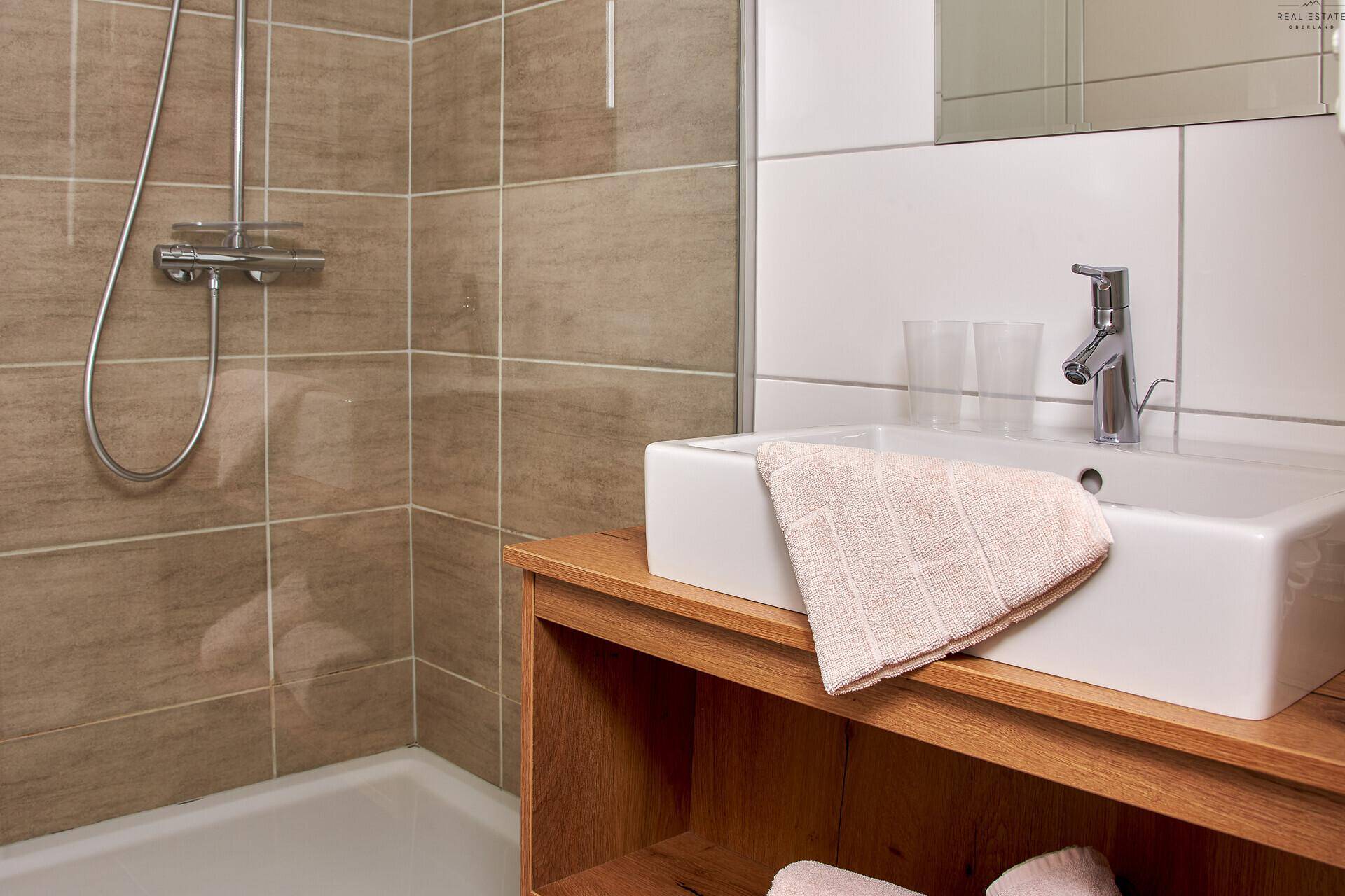 Modernes Badezimmer mit braunen Fliesen, Waschbecken auf Holzunterschrank und Duschbereich.