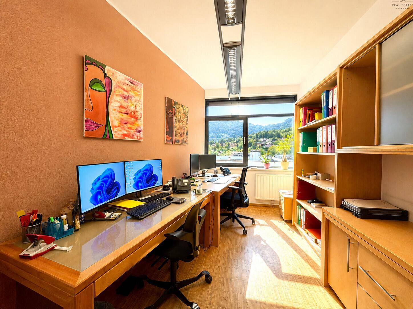 Bürozimmer mit zwei Arbeitsplätzen, mehreren Monitoren, Holzregalen und Blick ins Grüne.