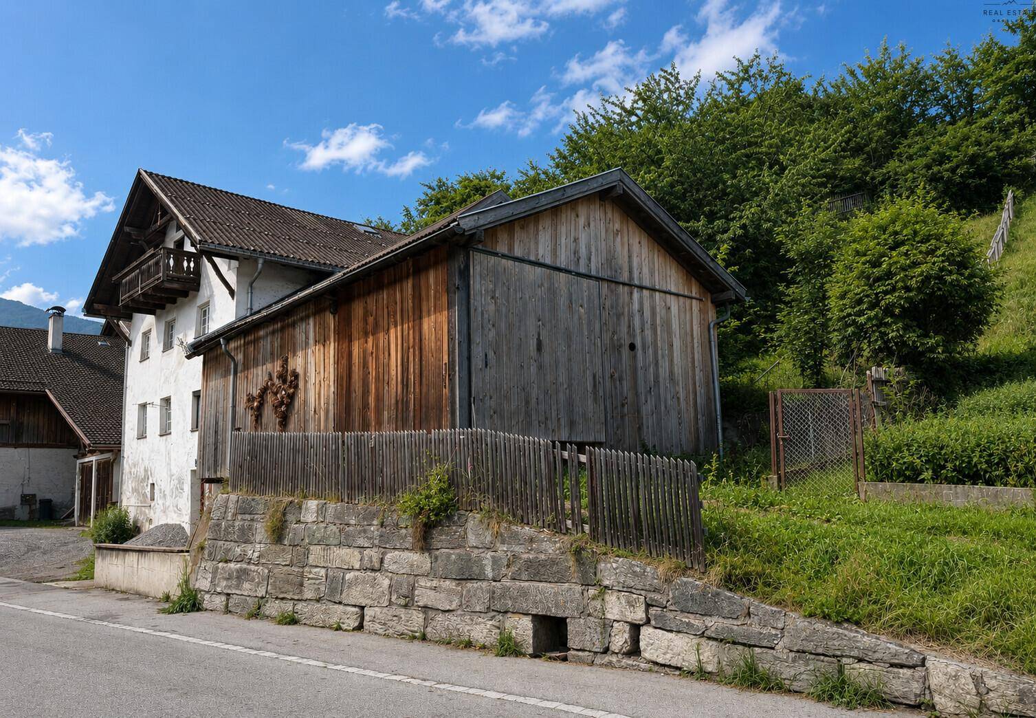 Traditionelles Bauernhaus mit Holzanbau und angrenzendem Hanggrundstück unter blauem Himmel.