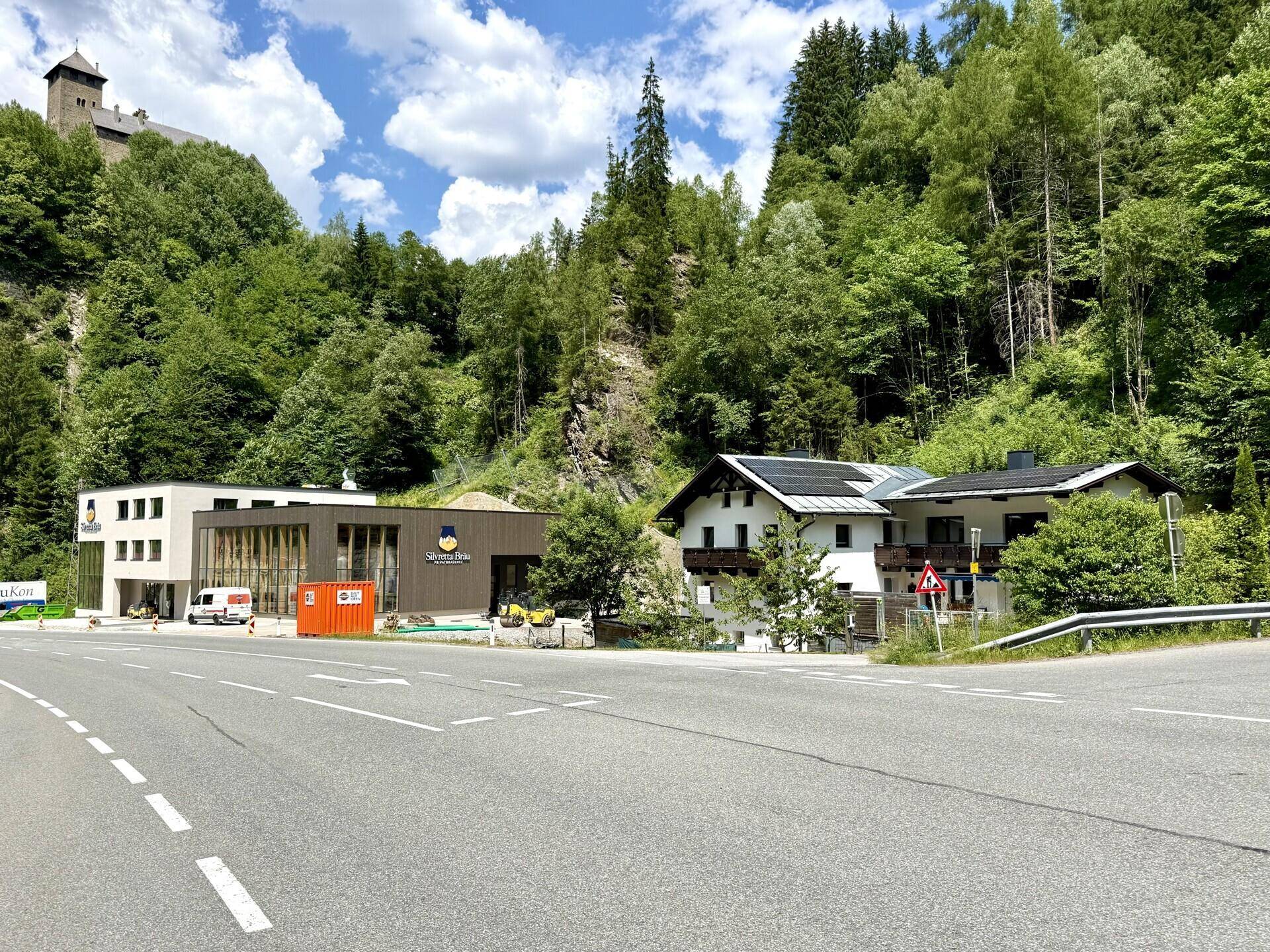 Gebäudeensemble in bergiger Landschaft mit Straße und Wald im Hintergrund.