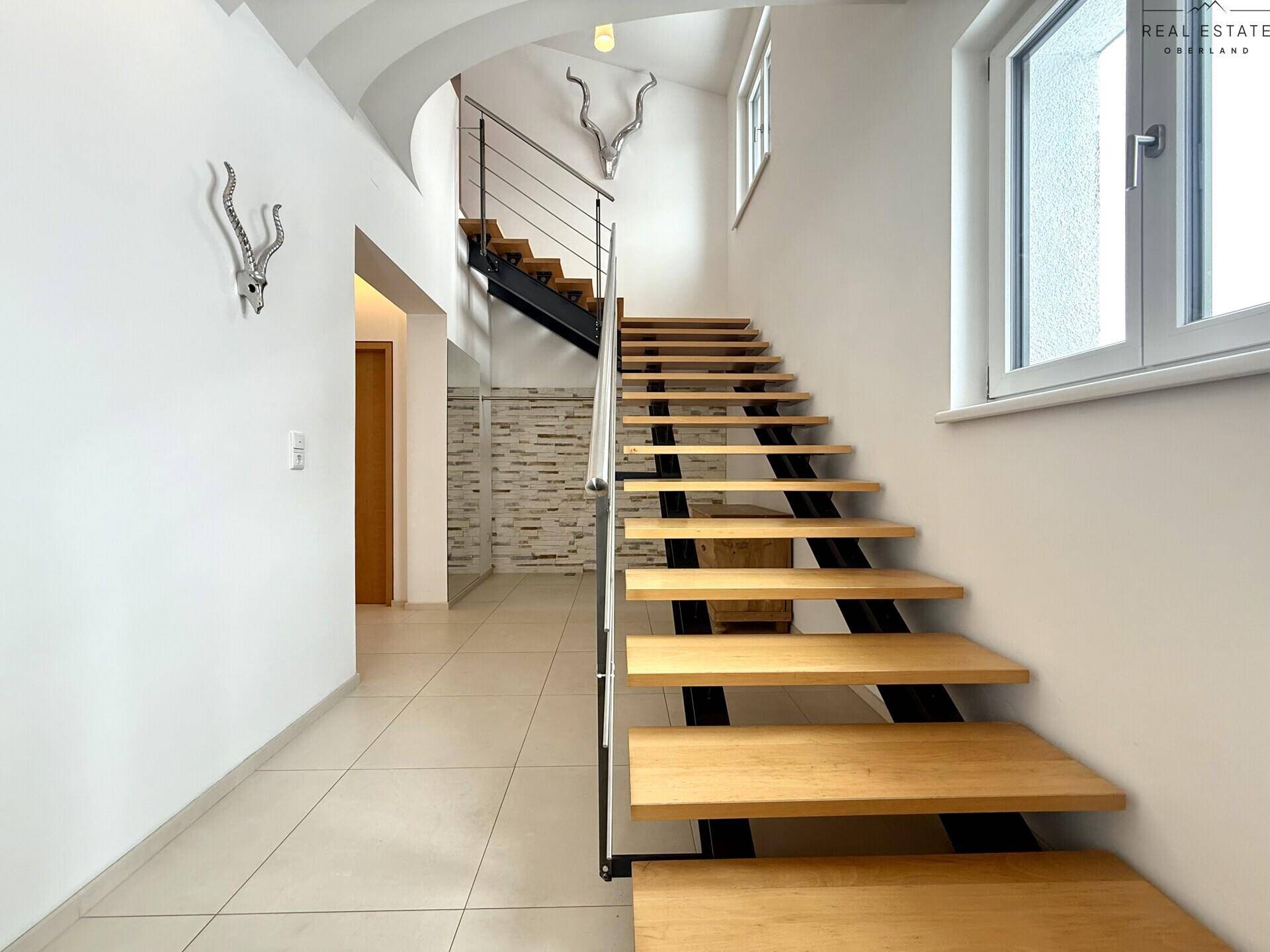 Moderne Holztreppe mit Metallgeländer und Fliesenboden im Eingangsbereich, führt in die oberen Etagen.
