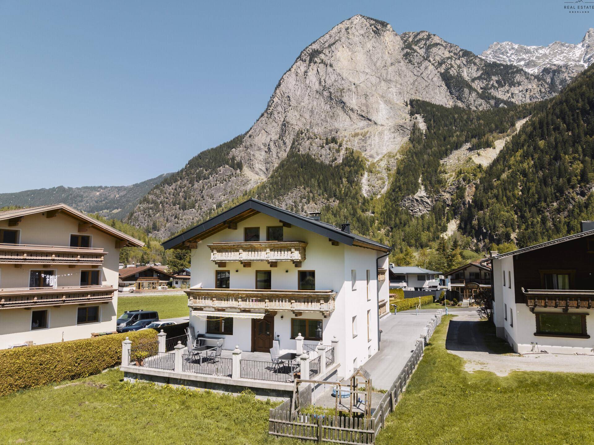 Traditionelles Haus mit Balkonen und Terrasse, umgeben von majestätischen Bergen und grüner Landschaft.