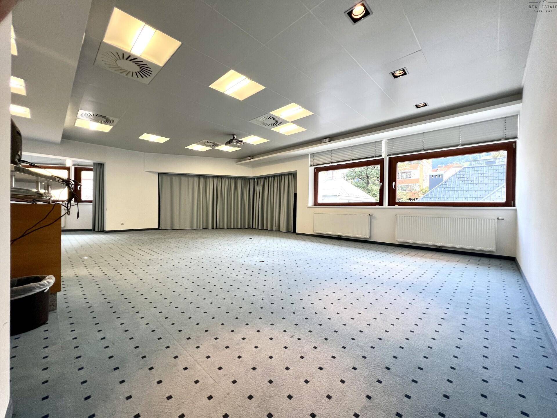 Großer, leerer Konferenzraum mit Teppichboden, Vorhängen und Fenstern mit Blick nach draußen.