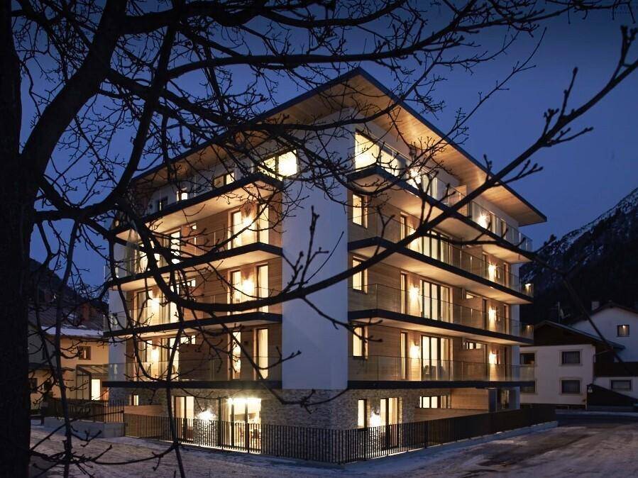 Modernes Apartmentgebäude bei Nacht, beleuchtet und umgeben von winterlicher Landschaft.