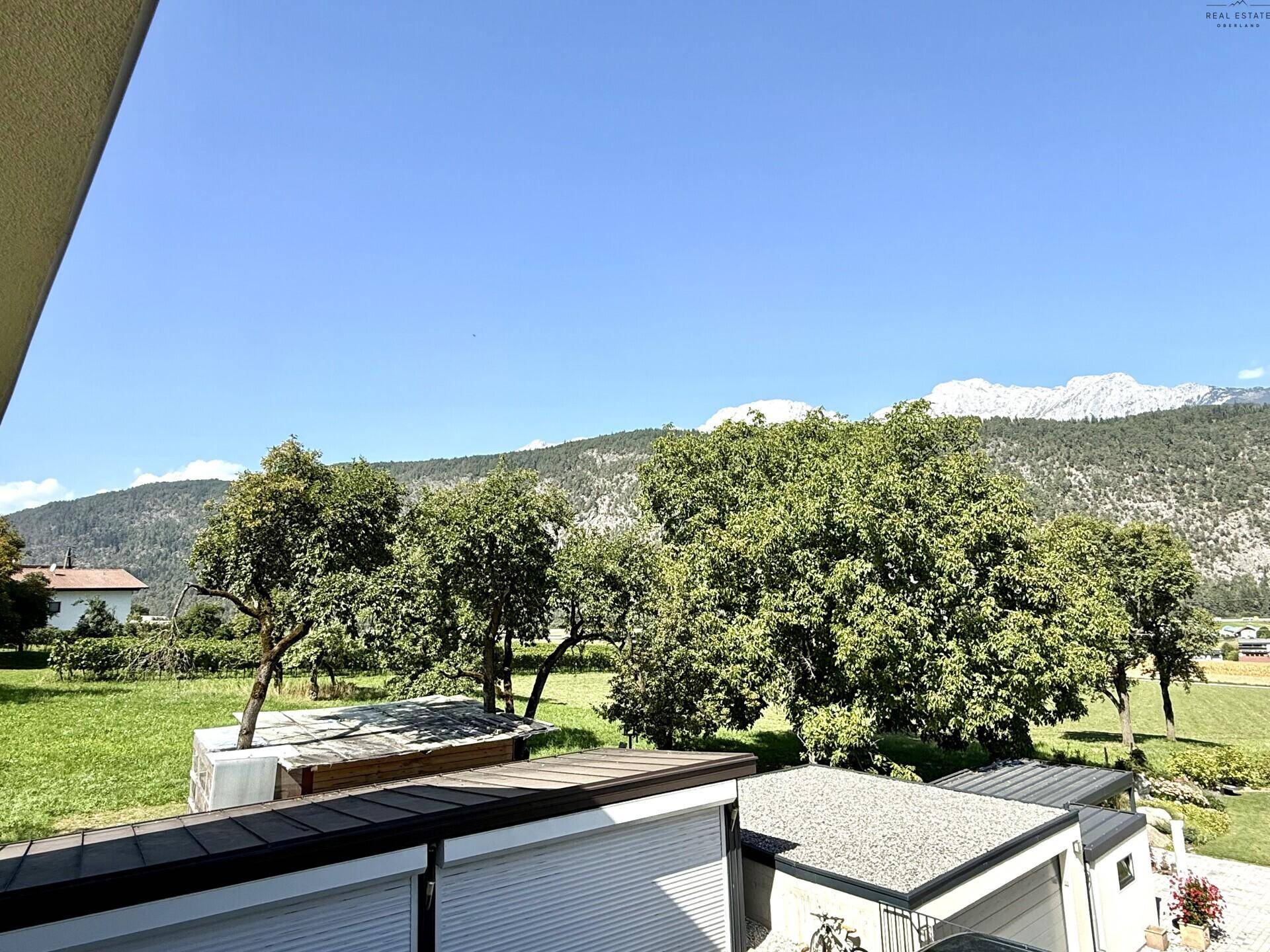 Panoramablick vom Balkon auf grüne Wiesen, Bäume und schneebedeckte Berge im Hintergrund.