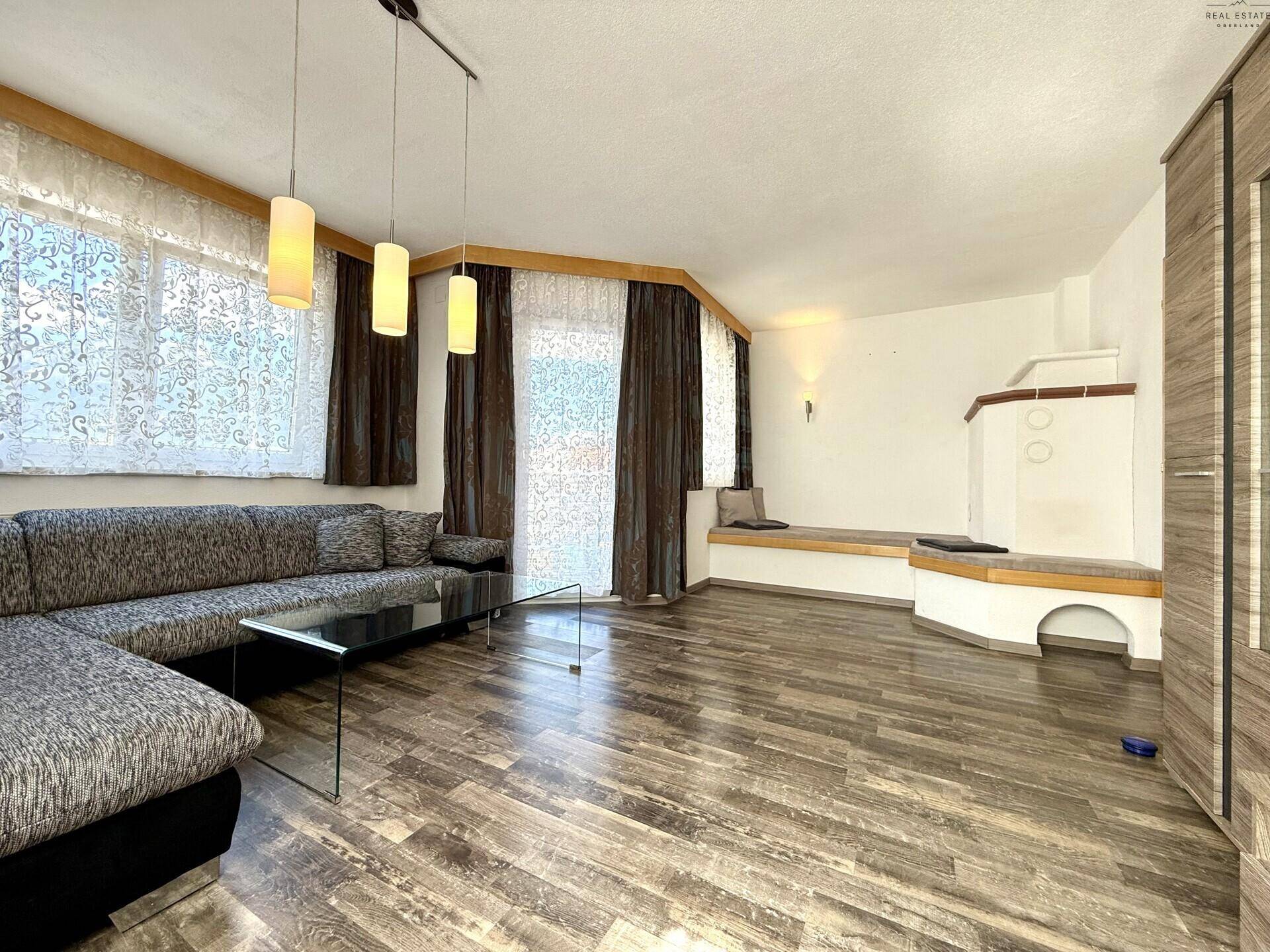 Gemütliches Wohnzimmer mit Ecksofa, Holzboden und traditionellem Kachelofen.